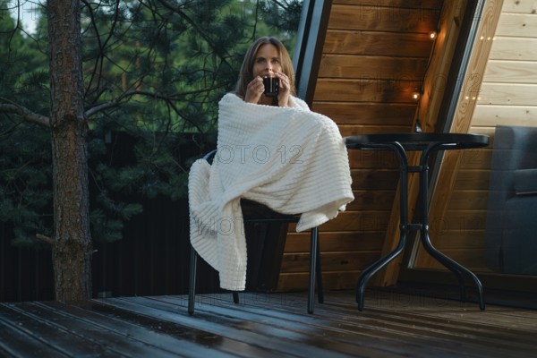 A woman relaxes on a chair while wrapped in a blanket. She holds a drink and enjoys fresh air after the rain. The setting is a wooden deck of tiny house surrounded by trees during the evening
