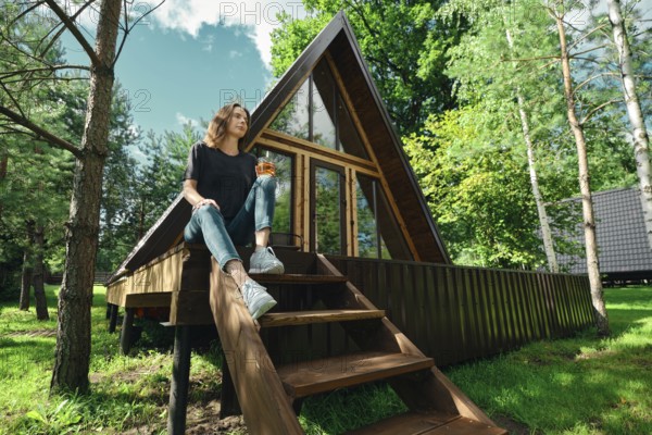 A woman sits on the steps of a wooden cabin surrounded by trees. She holds a drink and looks relaxed. The cabin has large windows and is in a sunny area. It is a calm day in the forest
