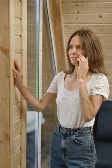 A woman stands near a wooden wall in a modern room while talking on her phone. She appears focused and engaged in conversation. The setting has large windows that let in natural light