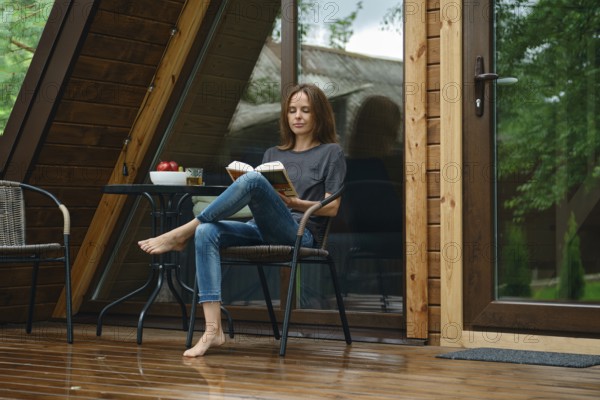 Barefoot woman sits on a chair on a wet wooden deck of tiny house after the rain. She is reading a book while enjoying apples placed on a small table. Green trees surround the area, and the sun is shining