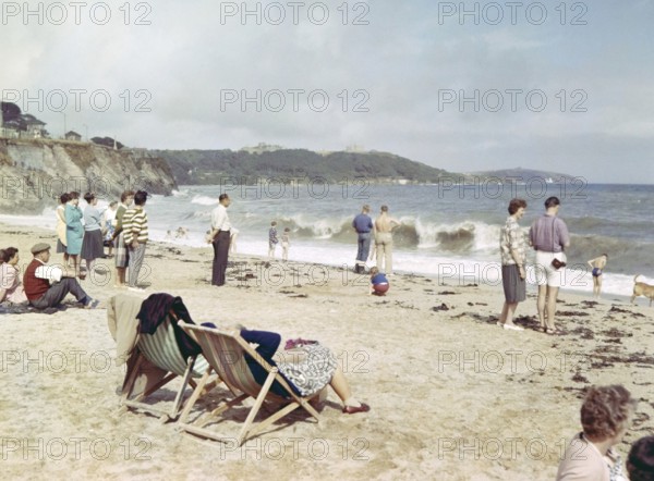 People on sandy beach looking at breaking waves, Gyllyngase beach, Falmouth, Cornwall, England, UK c 1960