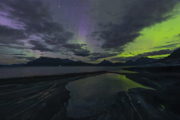 The Vestfjord in Nordland, Norway, has an enchanting atmosphere at night. Dark clouds and vivid aurora, polar lights dance over the fjord. The rugged cliffs and majestic mountains are reflected in the calm water and create a mystical backdrop