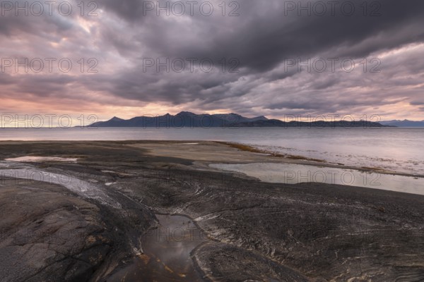 The Vestfjord in Nordland, Norway, offers a dramatic atmosphere near Bodø. Dark clouds are illuminated by the setting sun in shades of red and yellow. The rugged cliffs and mountains are reflected in the quiet fjord, which offers a breathtaking view of Lofoten