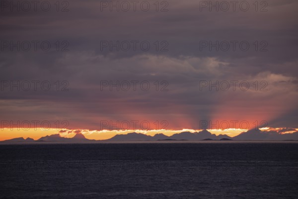 The Vestfjord in Nordland, Norway, offers a dramatic atmosphere near Bodø. Dark clouds are illuminated by the setting sun in shades of red and yellow. View towards Lofoten