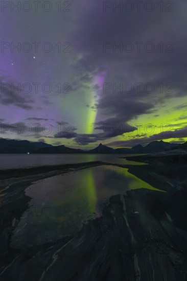 The Vestfjord in Nordland, Norway, has an enchanting atmosphere at night. Dark clouds and vivid aurora, polar lights dance over the fjord. The rugged cliffs and majestic mountains are reflected in the calm water and create a mystical backdrop