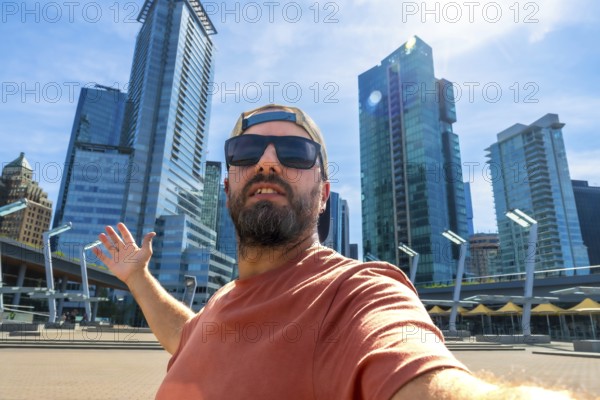 Bearded tourist wearing sunglasses and baseball cap takes selfie with outstretched arm in front of modern skyscrapers in vancouver, british columbia, enjoying sunny summer day