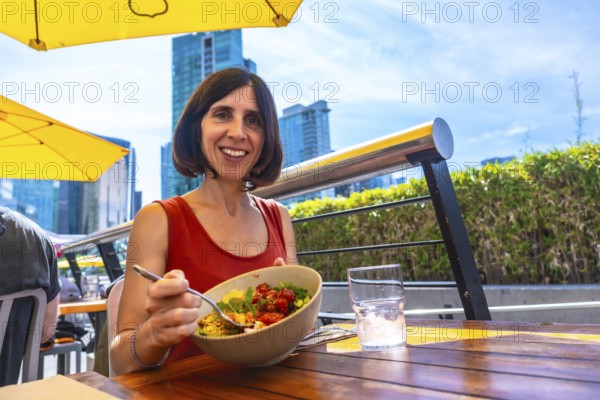 Happy woman enjoying a healthy poke bowl at an outdoor restaurant patio in vancouver, british columbia, with city skyline in background on a sunny summer day