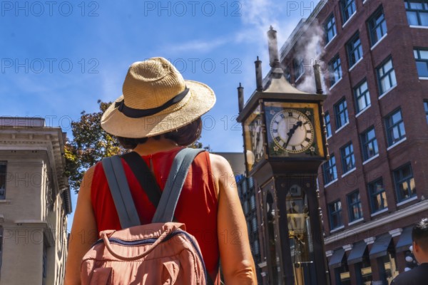 Female tourist wearing straw hat and backpack looking at the famous steam clock in gastown historic district, vancouver, british columbia, canada, in a sunny summer day