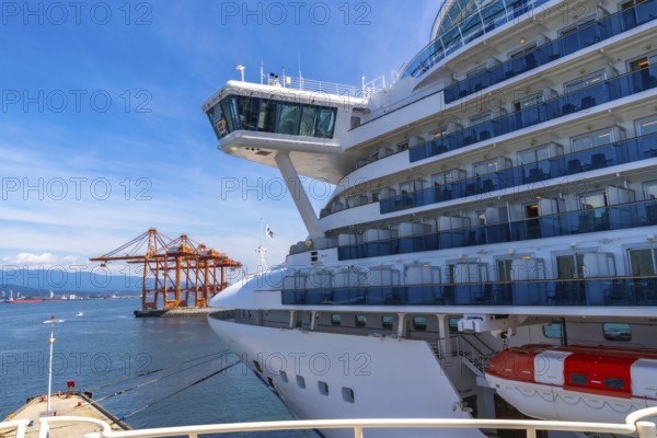 Large white cruise ship docking in vancouver harbor, surrounded by towering cargo cranes and majestic mountains under a bright blue sky on a sunny day, creating a picturesque scene