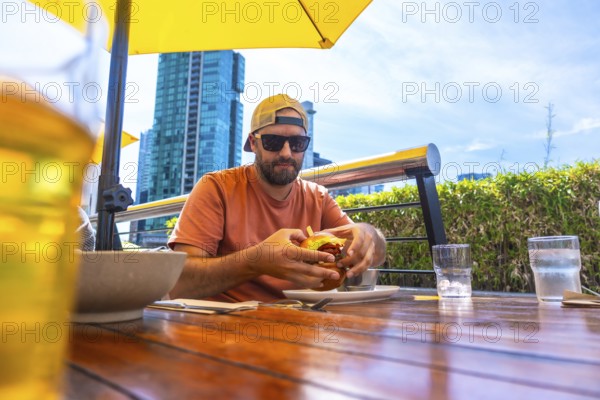 Tourist wearing sunglasses and a baseball cap enjoys a burger and beer on an outdoor patio restaurant in vancouver, british columbia, with city skyline in background
