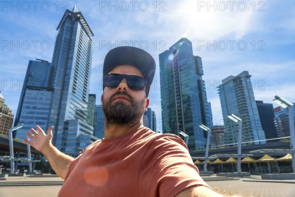 Happy bearded tourist wearing sunglasses and a baseball cap is taking a selfie with outstretched arms in front of modern skyscrapers in vancouver, british columbia, canada, on a sunny day