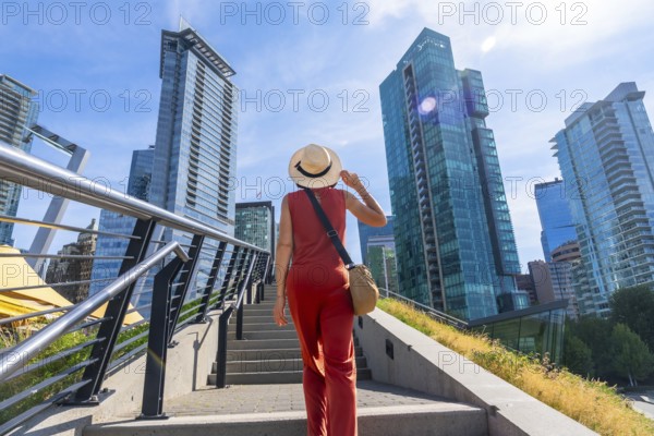 Female tourist wearing a straw hat and holding her hat is walking up stairs with modern glass skyscrapers in the background in vancouver, british columbia, canada