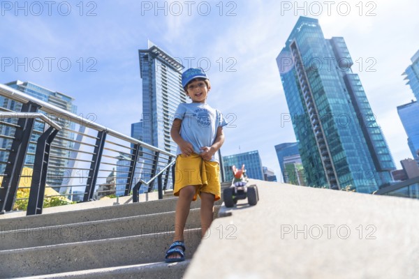 Young boy standing on steps playing with a toy car, with vancouver's modern buildings and clear blue sky as a backdrop, enjoying a sunny day in the city