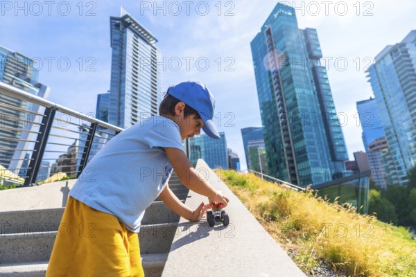 Young boy playing joyfully with a toy car on the steps of an urban park, soaking up the warmth of a sunny day, with vancouver's impressive skyline rising in the background