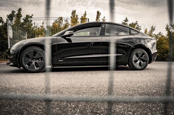 Side view of a black car in front of a fence surrounded by autumn leaves, Deer E- car sharing, Tesla Model 3, Calw, Germany