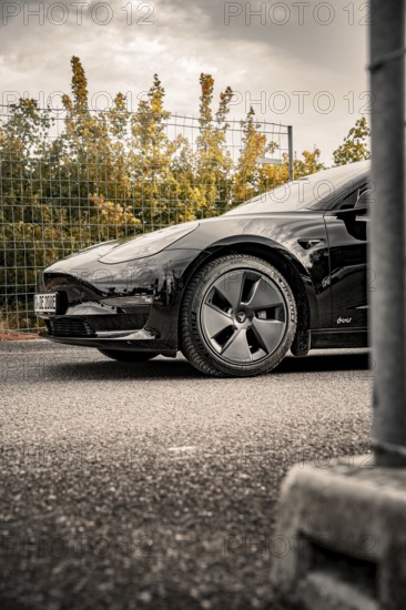 Black car on asphalt in front of a fence surrounded by trees and clouds, Deer E-Carsharing, Tesla Model 3, Calw, Germany