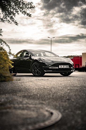 Black car in a parking lot under cloudy sky with trees nearby, Deer E- car sharing, Tesla Model 3, Calw, Germany