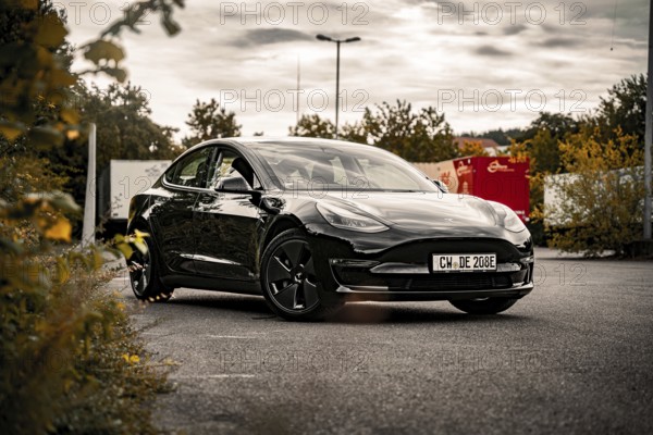 Black car on asphalt surface with cloudy sky and autumn vegetation, Deer E-Carsharing, Tesla Model 3, Calw, Germany