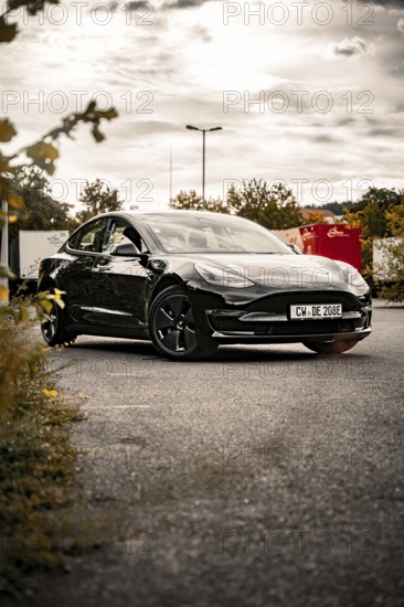 Black car on asphalt surrounded by grass and flowers with cloudy sky in the background, Deer e-Carsharing, Tesla Model 3, Calw, Germany