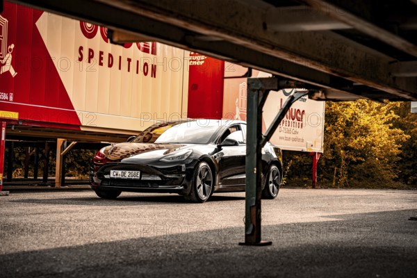 Black car parked next to freight forwarding trailers under a bridge-like structure, autumn trees in the background, deer e-car sharing, Tesla Model 3, Calw, Germany