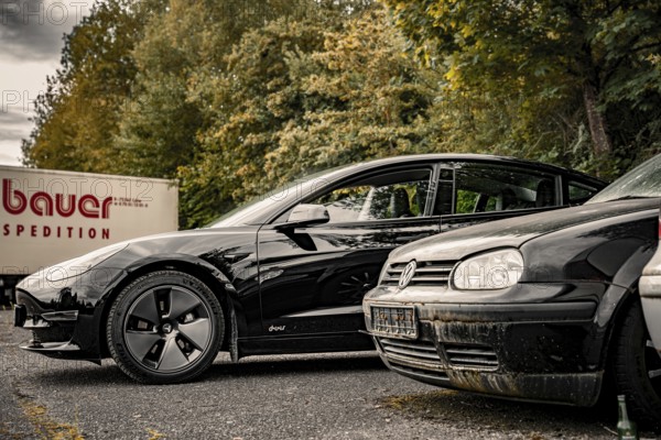 Black car and older vehicle in a parking lot with a truck in the background, Deer E-Carsharing, Tesla Model 3, Calw, Germany