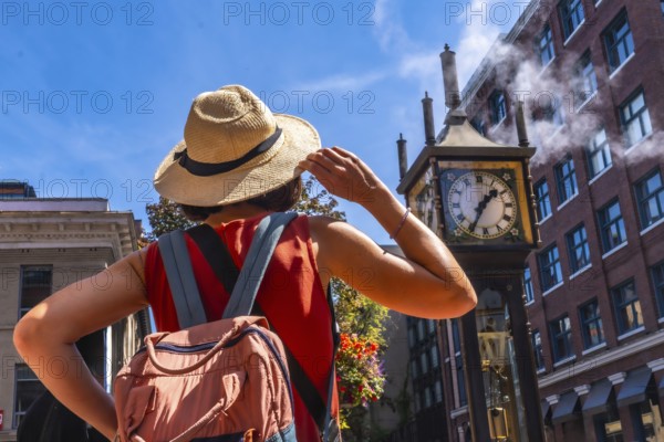 Young woman with backpack and straw hat looking at the famous steam clock in gastown, vancouver, enjoying a sunny summer day exploring the historic district of british columbia