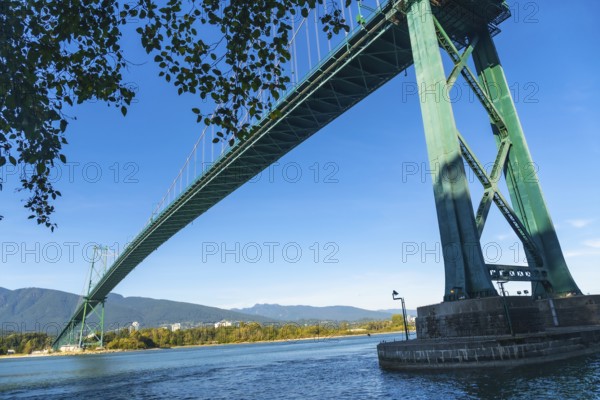Lions gate bridge extending over burrard inlet with stanley park seawall in foreground and north shore mountains in background on clear sunny day in vancouver, british columbia