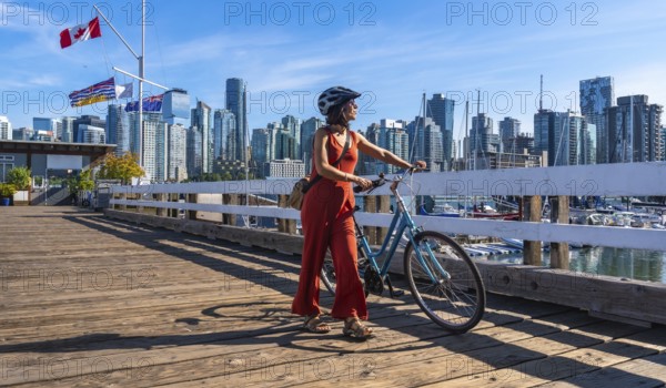 Female tourist wearing a helmet is walking alongside her bicycle on a wooden pier, enjoying the vibrant cityscape of vancouver against a backdrop of skyscrapers on a sunny summer day