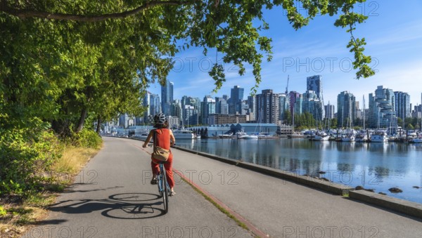 On a sunny summer day, a woman enjoys a leisurely bike ride along the seawall in stanley park, with the stunning cityscape of vancouver, british columbia, as a backdrop