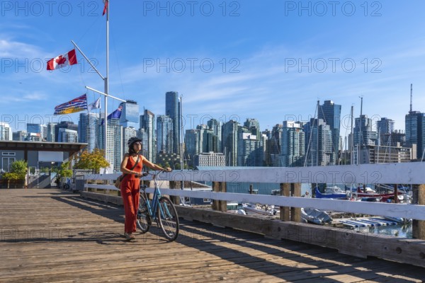 Tourist walking with her bicycle on a wooden pier with canadian and british columbian flags waving in the background, showcasing the vancouver skyline and harbor on a sunny day