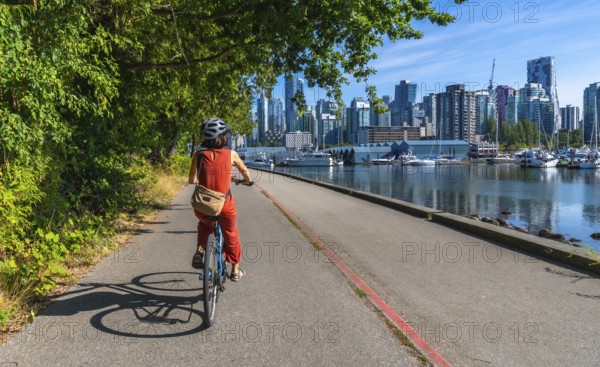 Tourist wearing helmet and orange jumpsuit is riding bicycle on seawall in stanley park with vancouver city skyline, marina and sea in background on sunny summer day