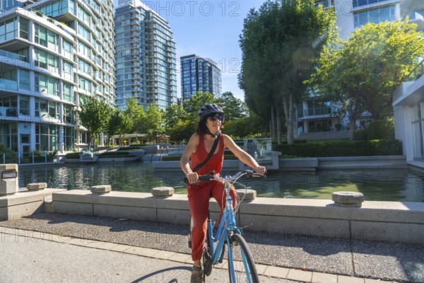 Woman wearing a helmet and sunglasses is riding a bicycle along a waterfront path in vancouver, british columbia, enjoying the cityscape and sunny weather