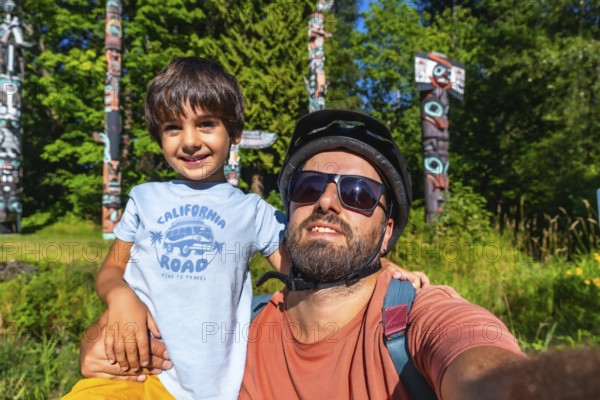 Father and son enjoying a sunny day at brockton point in stanley park, vancouver, taking a selfie with traditional totem poles in the background