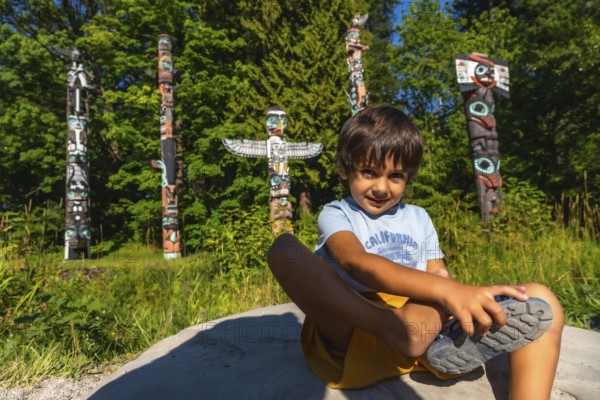 Young boy sitting on a rock, tying his shoe in front of vibrant totem poles at brockton point in stanley park, vancouver, british columbia, enjoying a sunny day outdoors