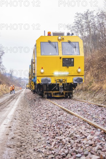 Yellow rail vehicle travels on gravel tracks through wintry landscape with workers, stuffing machine on Hermann, Hesse, railway, Althengstett, Calw district, Germany