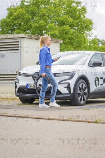 Woman standing on the street with a white electric car with charging cable, Deer E-Carsharing, Calw, Germany