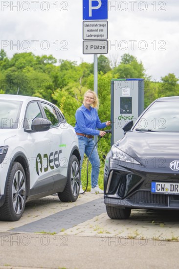 Woman refuels electric car at a charging station between two parked vehicles, Deer E-Carsharing, Calw, Germany