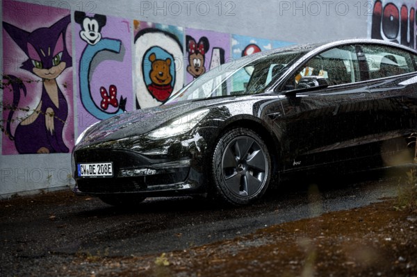 Black car in front of a rain-covered road and colorful graffiti wall, Deer E-Carsharing, Tesla Model 3, Calw, Germany
