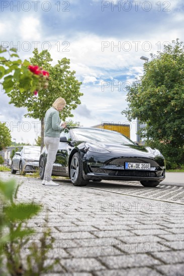Person standing next to black car on paved road under blue sky with clouds, Deer E-Carsharing, Tesla Model 3, Calw, Germany