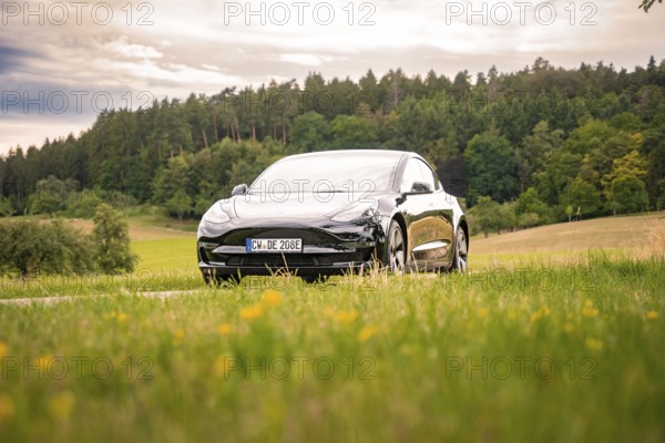 Black car on a country road surrounded by meadows and forest under cloudy sky, Deer E-Carsharing, Tesla Model 3, Calw, Germany