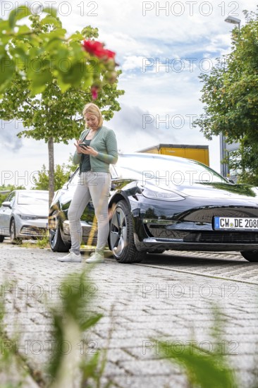 Woman using a tablet next to a parked black car outdoors, Deer E- car sharing, Tesla Model 3, Calw, Germany