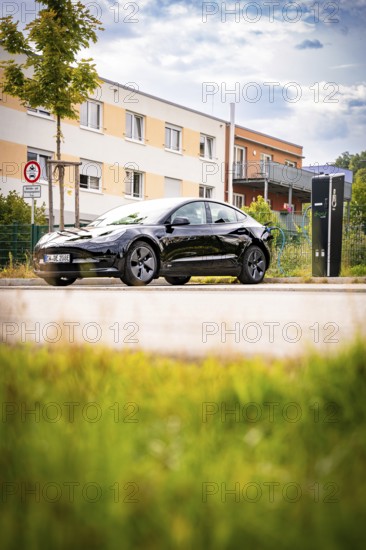 Black car at a charging station in front of a modern building backdrop under a blue sky, Deer E-Carsharing, Tesla Model 3, Calw, Germany