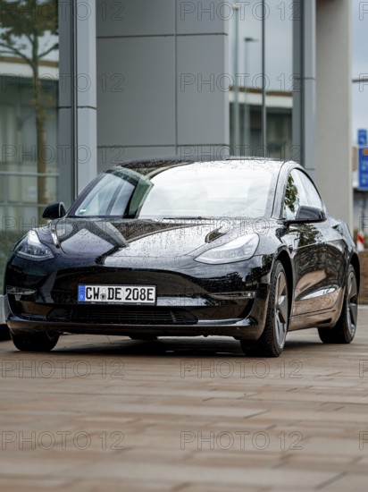 Black car with rain droplets on paint in front of glass buildings, Deer E-Carsharing, Tesla Model 3, Calw, Germany