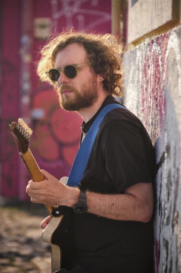 Hipster street musician in black playing electric guitar in the street on sunset leaning on a wall. Lisbon, Portugal
