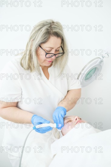 Cosmetologist in blue gloves delivers a hydrafacial cleansing treatment to a client, using blue light for skin rejuvenation