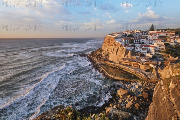 The picturesque village of Azenhas do Mar is illuminated during sunset, resting on cliffs above the Atlantic Ocean. Waves crash below, creating a stunning coastal scene. Portugal