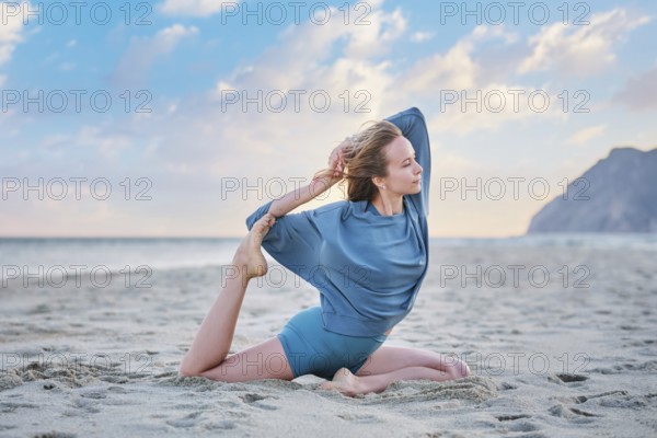 A woman practices Eka Pada Rajakapotasana on the sand during sunset, focusing on her yoga pose with the ocean in the background