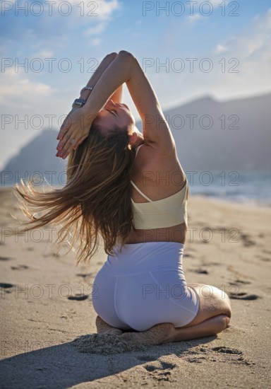 Woman performs Vajrasana with backbend on sandy beach as the sun sets over the ocean. She kneels on the sand with arms raised