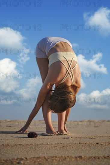 A woman performs Uttanasana on a sandy beach during sunset while clouds fill the sky. She leans forward and touches the sand
