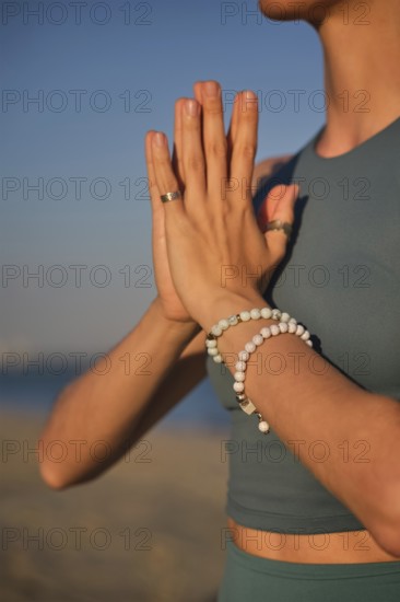 A woman practices yoga by holding hands in Namaskar Mudra at the beach as the sun sets in the background, creating a stunning scene